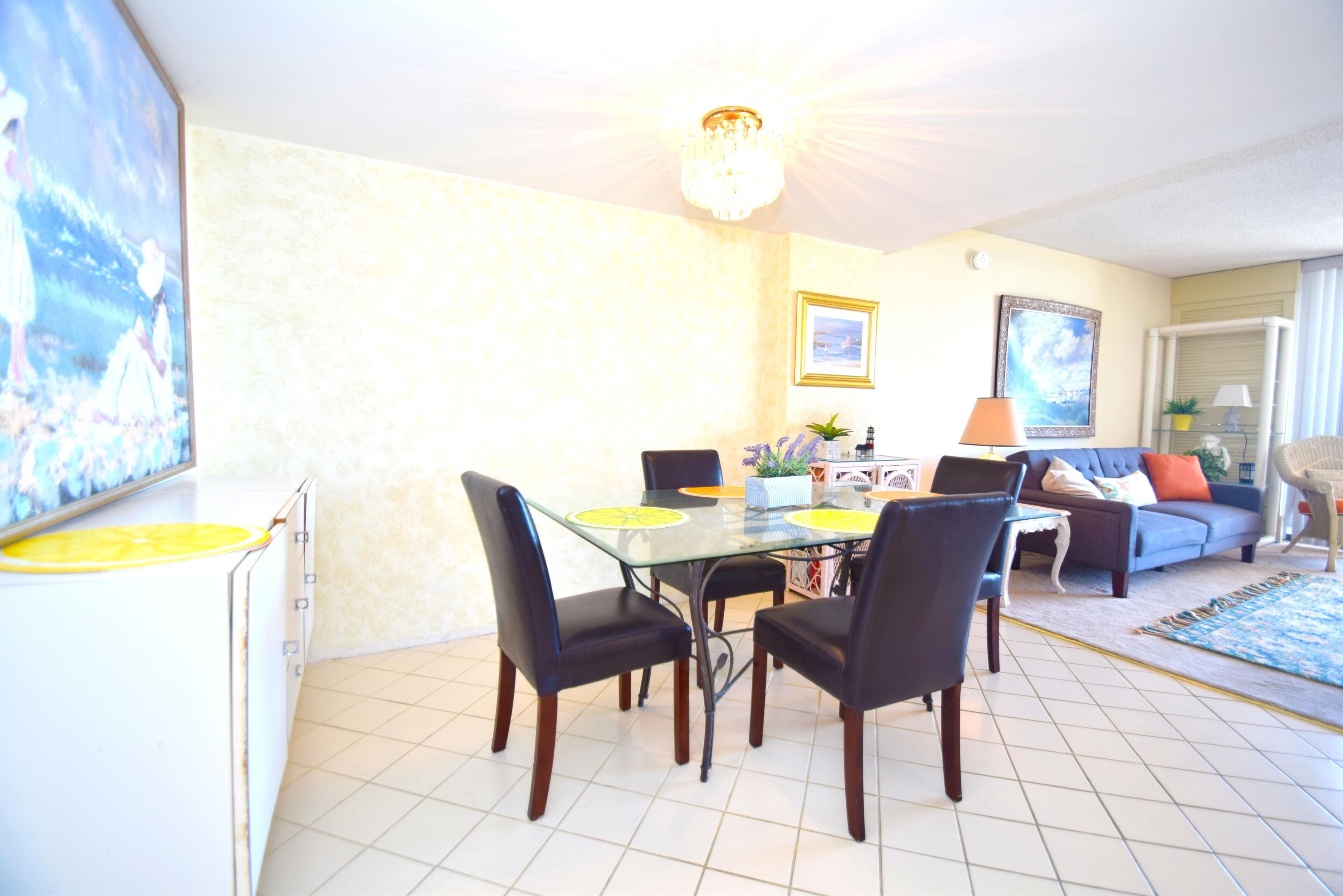 Un coin repas avec une table en verre, quatre chaises, un lustre et un canapé en arrière-plan.