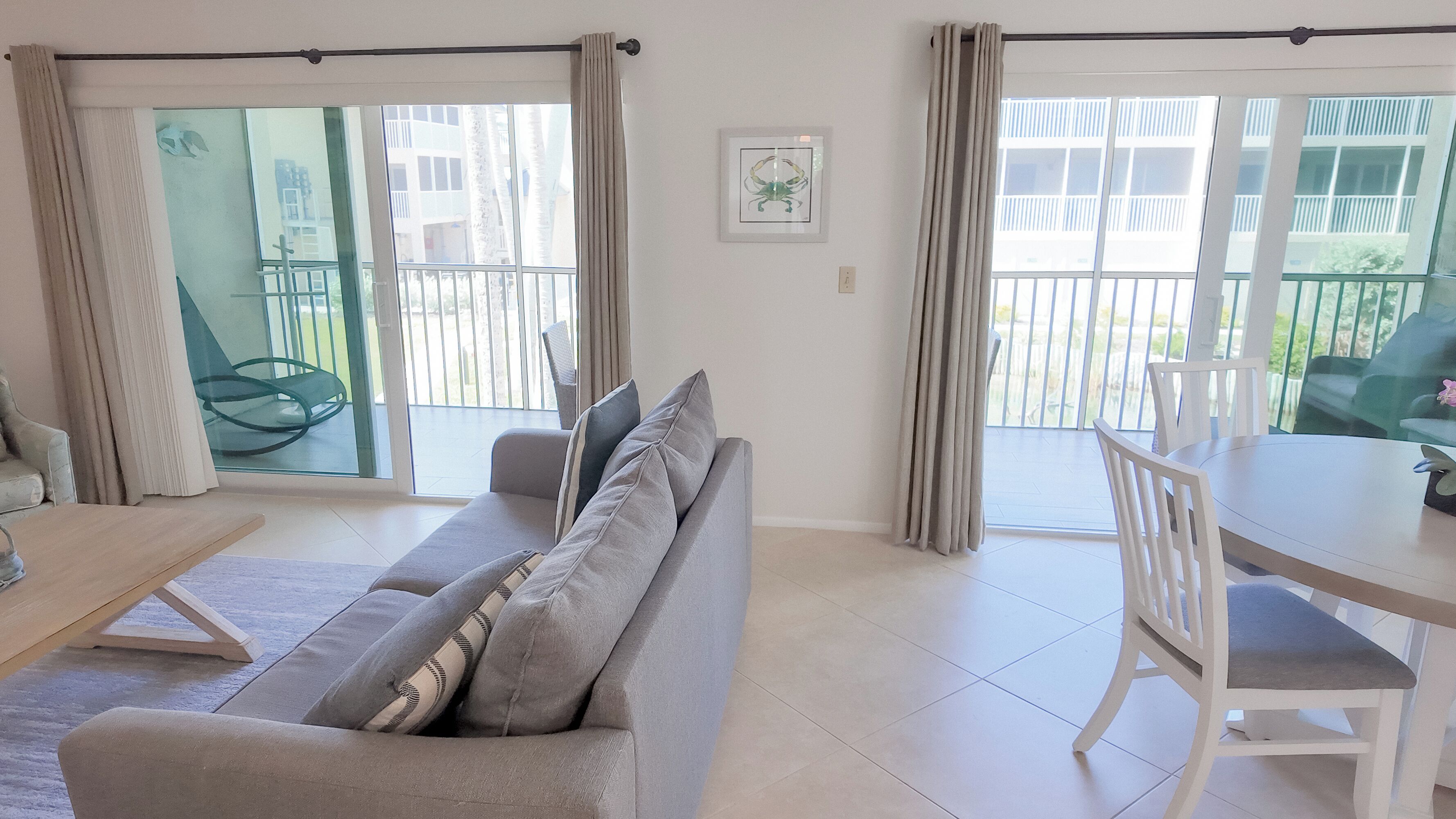 A modern living room with a grey sofa, a wooden coffee table, and a white dining table with chairs. There is a sliding glass door leading to an outdoor balcony.
