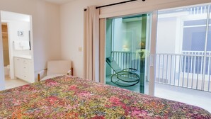 A bedroom with a floral bedspread, a chair, a balcony with a glass door, and a white bathroom with a towel rack. - Serene and Relaxing (Sanibel)