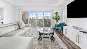 A modern living room with a glass coffee table, white sofa, teal armchair, and a large TV. - Sunset View | Stunning Golf Views Close to Disney! Luxury Condo Near Disney! (Kissimmee)