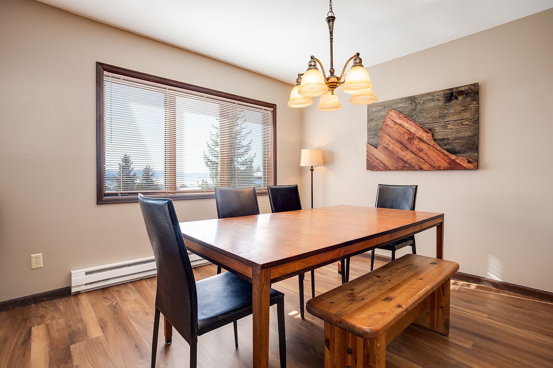 A dining area with a wooden table, four chairs, a bench, a chandelier, a floor lamp, and a large window with blinds.