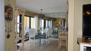 A dining area with white chairs and a glass table, a television on a stand, and a large mirror reflecting the room.