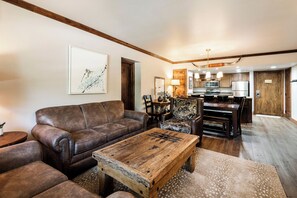 A living room with a brown leather sofa, a wooden coffee table, and a patterned armchair. There is a kitchen in the background with modern appliances.