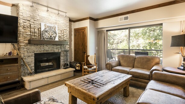 A cozy living room with a stone fireplace, leather sofas, and a large window offering a view of trees.