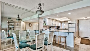 A modern kitchen with white cabinets, a central island, and a dining area with white chairs and a glass table. - El Matador 413 by Alicia Hollis Rentals FREE TICKETS! $300/Day (Fort Walton Beach)