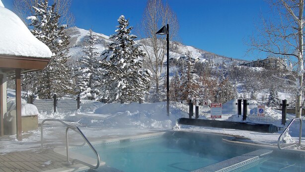 A heated outdoor pool with a metal railing, surrounded by snow-covered trees and mountains.