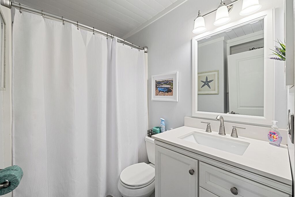A bathroom with a white shower curtain, a white toilet, a white sink with a mirror above it, and a framed picture on the wall.