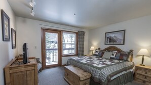 A bedroom with a large bed, a wooden chest, a desk with a computer, and a window with a view of trees.