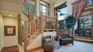 A spacious living area with a wooden staircase, a large window with a view of trees, and a cozy seating area with a patterned ottoman and chairs.