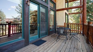 A wooden deck with a glass door entrance, a small outdoor table and chairs, and a view of a building with a balcony.