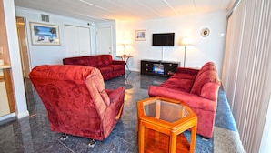 A living room with red sofas, a glass coffee table, a flat-screen TV, and a door leading to another room. - Thunder Island 10A-Baywater 39th St, Pool, Elev, W/D, AC (Ocean City)