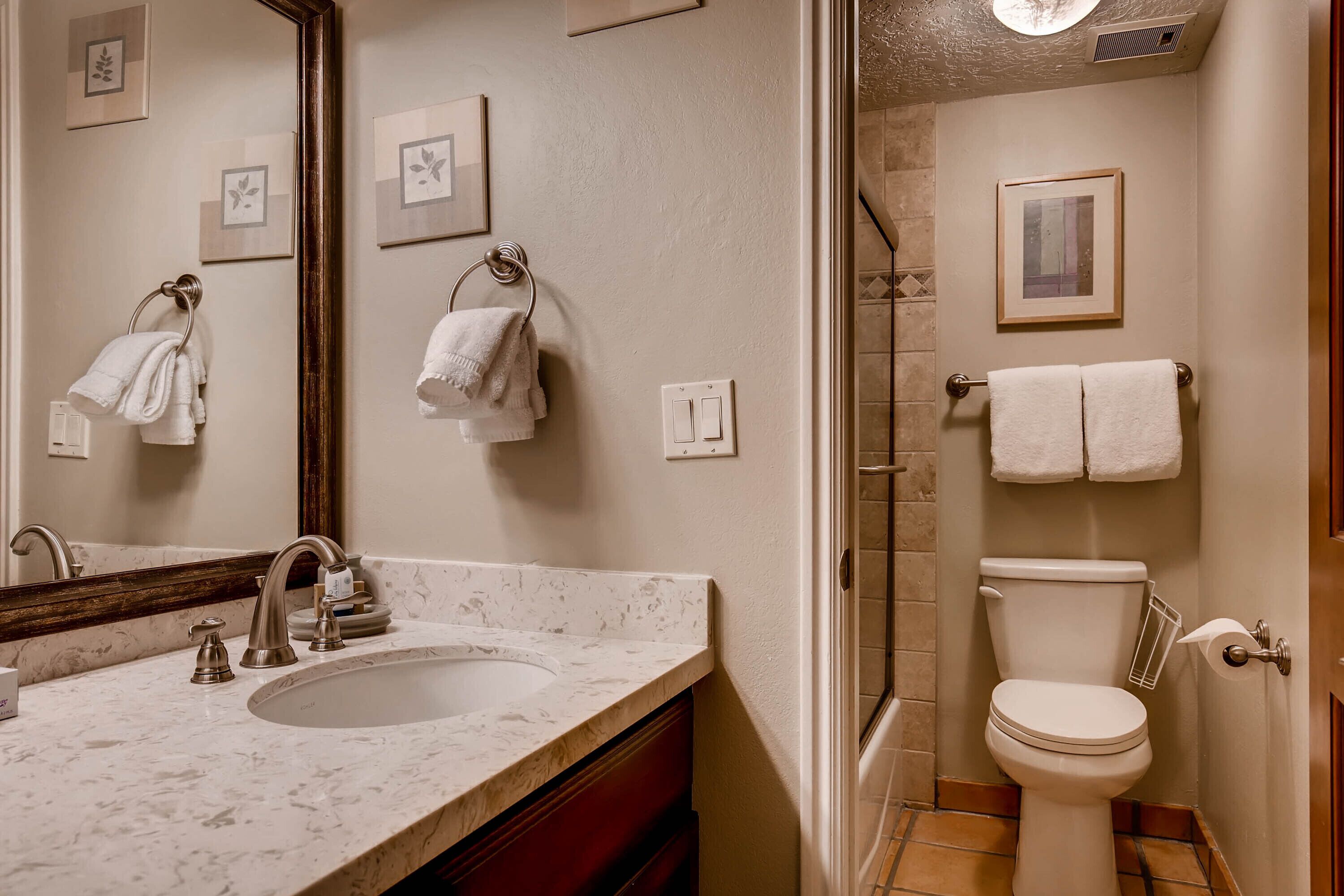 A bathroom with a sink, mirror, and toilet. There are towels hanging on the wall and a shower area visible.