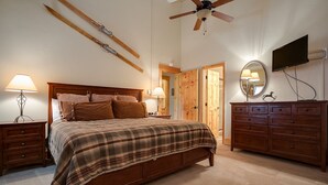 Une chambre à coucher avec un lit en bois, deux tables de chevet avec des lampes, une commode, un ventilateur de plafond et un téléviseur fixé au mur.