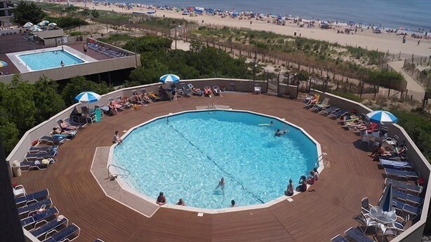 A circular pool surrounded by sun loungers and umbrellas, with a beach view in the background. - High Point South 3F-Oceanfront 112th St,Free WiFi,Pool,Elev,W/D,AC (Ocean City)