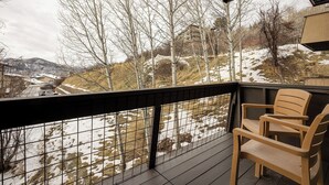 Ein Balkon mit einem Holzbstuhl, einem Metallgeländer und Blick auf eine verschneite Landschaft mit Bäumen und einem Gebäude in der Ferne.