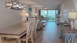 Amplia sala de estar con mesa de comedor de madera, sillas blancas, sofá y televisor. Hay un balcón con vistas al exterior.