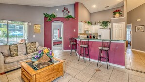 A spacious living room with a sofa, a coffee table, and a kitchen area with a bar and stools. - Enjoy In Pool Dining, Hot Tub & Games Rm. Disney 4 Miles. Pet Friendly (Kissimmee)