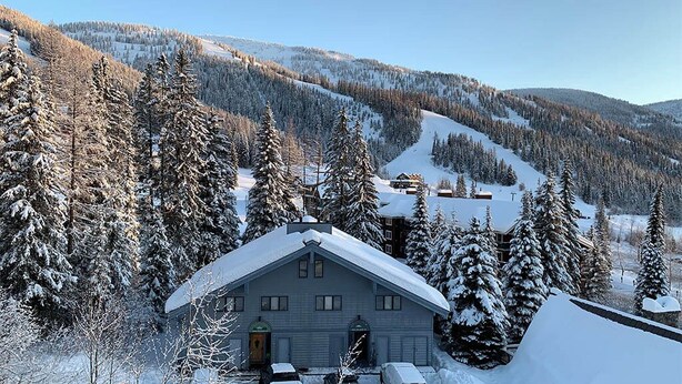 A snow-covered lodge with a garage parking area and a view of a snowy mountain landscape. - Condo with Excellent Mountain Views (Whitefish)
