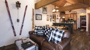 A cozy living area with a brown leather couch, a small table, and skis hanging on the wall. A kitchen is visible in the background with wooden cabinets and a microwave. - Condo with Excellent Mountain Views (Whitefish)