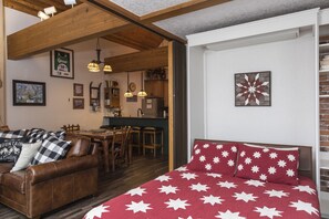 Quarto aconchegante com sofá de couro marrom, cama com padrão de estrelas vermelhas e brancas, vigas de madeira, área de jantar e cozinha ao fundo.