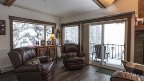 A spacious living room with leather sofas and a fireplace, large windows with a view of snow-covered trees, and a balcony with outdoor furniture. - Condo with Excellent Mountain Views (Whitefish)