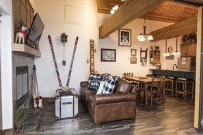 Uma sala de estar acolhedora com lareira, um sofá de couro marrom, uma mesa de jantar com cadeiras e uma cozinha com armários de madeira e luminárias pendentes.