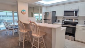 Een moderne keuken met witte kastjes, een roestvrijstalen koelkast, een magnetron en een oven. Er zijn twee houten barkrukken en een klein eiland met een spoelbak.