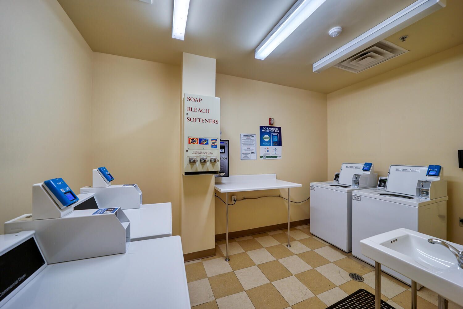 Une buanderie équipée d’un évier, de deux machines à laver, d’un sèche-linge et d’un distributeur de savon et d’eau de Javel.