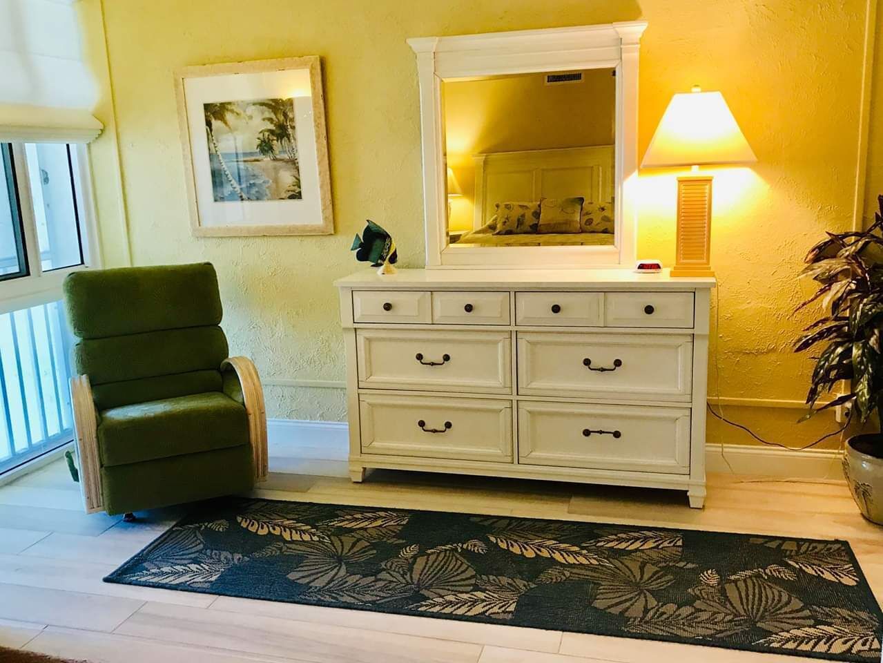 Una habitación con un sillón verde, un aparador blanco con cajones, un espejo, una lámpara y una planta en maceta.