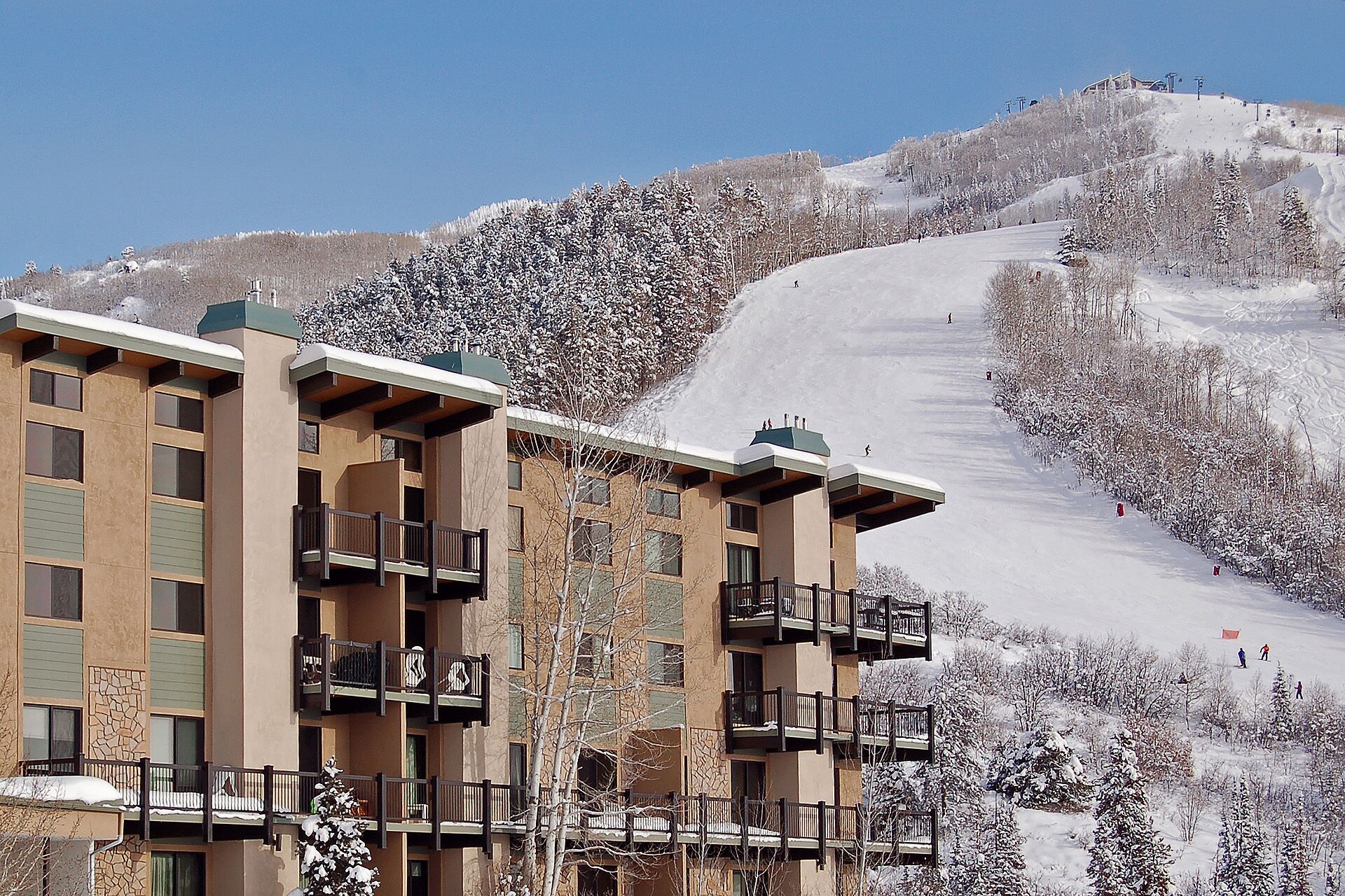 A multi-story hotel with balconies, a ski slope in the background, and snow-covered trees.