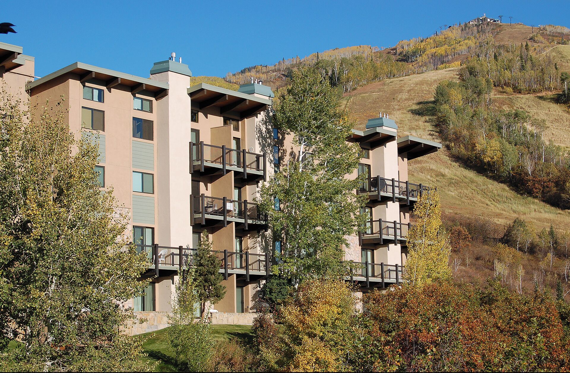 A multi-story hotel with balconies, set against a backdrop of a hilly landscape with autumn foliage.