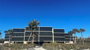 Un bâtiment moderne à plusieurs étages, doté d’un toit plat et de grandes fenêtres, entouré de palmiers et de petits arbustes.