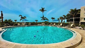 A circular pool with clear blue water, surrounded by lounge chairs and umbrellas, with palm trees and a building in the background. - Paradise Found at Atrium 203 Sanibel; 3br/3ba + Cabana (Sanibel)
