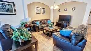 Una sala de estar con un sofá, dos sillones, una mesa de centro y un televisor. Hay plantas sobre la mesa de centro y una lámpara en la mesita auxiliar.