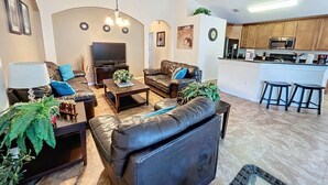 Una sala de estar con televisor, sofás de cuero, una mesa de centro y una cocina con microondas y taburetes.