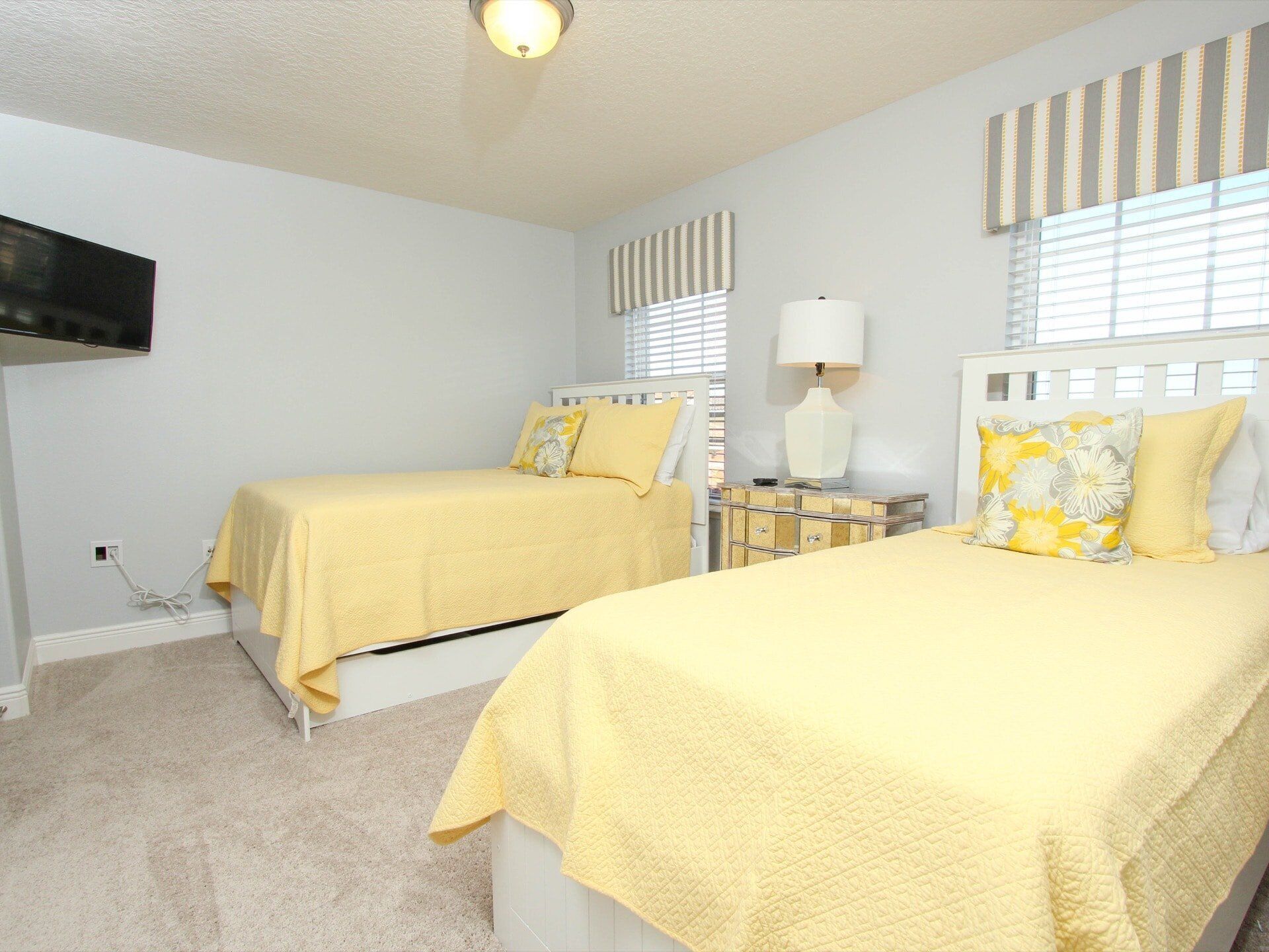 A bedroom with two beds, a bedside table with a lamp, a TV mounted on the wall, and a window with blinds.
