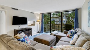Ein Wohnzimmer mit Flachbildfernseher, Lampe, Sofagarnitur und einem Balkon mit Blick auf Gebäude und Bäume.