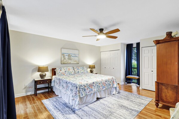 A bedroom with a bed, two bedside lamps, a ceiling fan, a painting, a wardrobe, and a window with curtains.