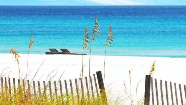 A beach with a wooden fence, a lounge chair, and tall grass in the foreground. - Surf Dweller 103 by Alicia Hollis Rental FREE ADVENTURES $300/Day (Fort Walton Beach)