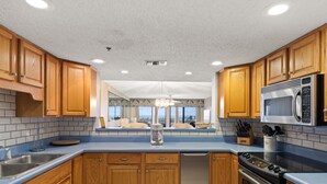 A modern kitchen with wooden cabinets, stainless steel appliances, and a large window offering a view. - Surf Dweller 103 by Alicia Hollis Rental FREE ADVENTURES $300/Day (Fort Walton Beach)