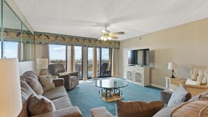 A spacious living room with a large glass coffee table, comfortable seating, a ceiling fan, and a view through large windows. - Surf Dweller 103 by Alicia Hollis Rental FREE ADVENTURES $300/Day (Fort Walton Beach)