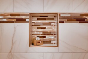 Un baño con pared de mármol y estante empotrado con artículos de aseo.