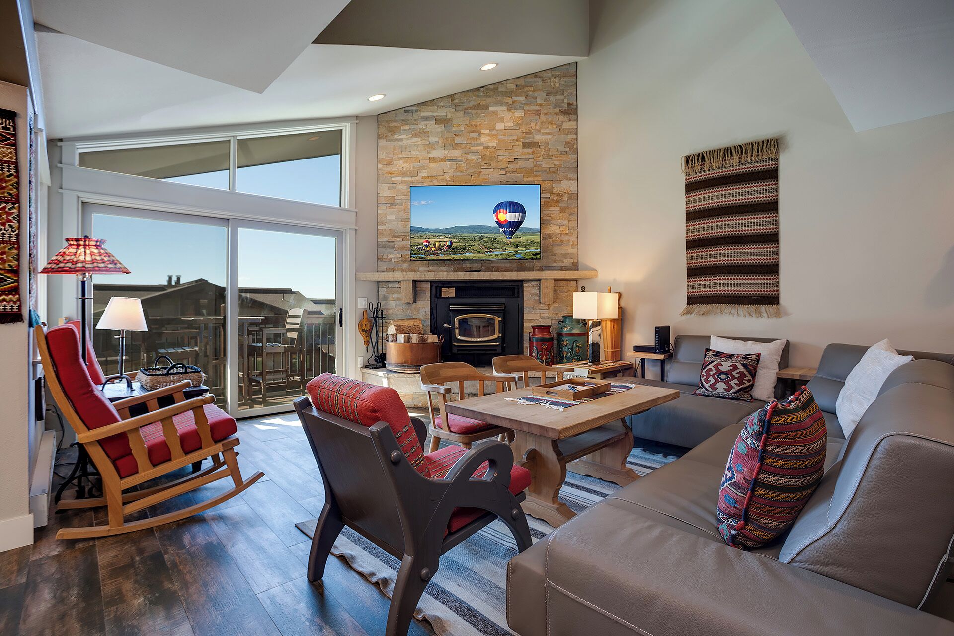 A spacious living room with a stone fireplace, a television mounted above it, and a large sliding glass door leading to a balcony.
