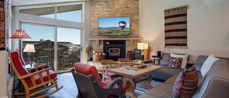 A spacious living room with a stone fireplace, a television mounted above it, and a large sliding glass door leading to a balcony.
