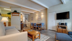Ein modernes Wohnzimmer mit Sofa, Couchtisch und Fernseher. Es gibt eine Küchenzeile mit gelben Schränken und einem Kühlschrank.