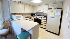 A compact kitchen with white cabinets, a microwave, a stove, a sink, and a refrigerator. There is a wooden chair with a cushion and a small table with a basket on top. - Ocean Princess 207-Oceanside 134th St, Elev, W/D, AC (Ocean City)