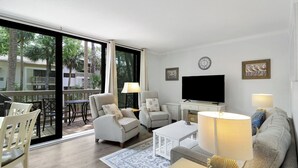 Uma sala de estar com sofá, poltronas, mesa de centro, televisão e varanda com mobiliário de exterior.