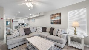 Un salon moderne avec un canapé d’angle, une table basse en bois, un ventilateur de plafond et une cuisine en arrière-plan.