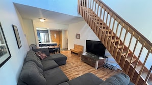 A living room with a sectional sofa, a TV on a wooden stand, a dining area with a table and chairs, and a wooden staircase. - East Village Walk To Super Bee Lift Restaurants and Shops Hot Tub Access (Copper)