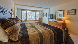 A bedroom with a large bed, a dresser, a mirror, and a window with a view of snow-covered trees. - Summit House Short Walk to Skiing, Covered Parking Balcony w/ Panoramic Views (Copper Mountain)